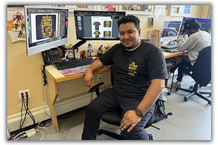 Person sitting at a desk with dual monitors displaying a design on one screen; another person working at a computer in the background; creative office setting.